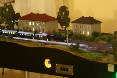 Model parowego pociągu pasażerskiego przemierza makietę krajobrazu, obok stoją modele budynków o prostej, europejskiej architekturze. Scena modelarska jest oświetlona tak, by imitować zmrok lub wczesny wieczór, co widać po oświetleniu budynków i 