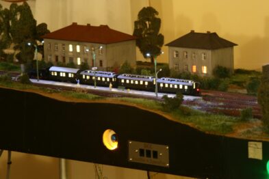 Model pociągu pasażerskiego z kilkoma wagonami przemierza makiety kolejowej, na tle modeli budynków, które wyglądają jak mieszkalne domy oraz drzewa. Model jezdni jest zarysowany na pierwszym planie, a tory kolejowe biegną przez środek sceny. Oświetlenie makiet budynków i latarni nadaje scenie realistyczne uczucie zmierzchu lub wczesnego wieczoru.
