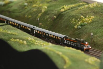 Model pociągu jedzie przez ukształtowany teren z zielonymi wzgórzami i niewielkimi elementami roślinności. Stworzony krajobraz jest szczegółowy, z uwzględnieniem elementów takich jak drobne rośliny i trawki. Lokomotywa wraz z wagonami ma barwy czarno-czerwone i symuluje ruch na miniaturowych szynach.