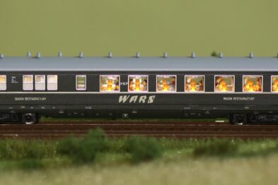 Model kolejki w skali TT przedstawia szczegółowo odwzorowany wagon pasażerski w kolorze granatowym z białym pasem. Okna są przejrzyste, przez co widać siedzących pasażerów i wnętrze wagonu. Wagon jest oznaczony białymi literami 