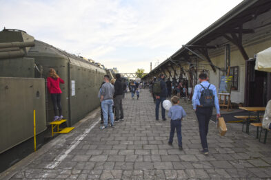 Odwiedzający spacerują po zewnętrznej przestrzeni muzeum kolejnictwa, gdzie ustawione są historyczne wagony kolejowe. Ludzie różnych grup wiekowych, w tym rodziny z dziećmi, korzystają z ciepłego dnia, aby zwiedzać i uczestniczyć w wydarzeniu. Na pierwszym planie widać osobę robiącą zdjęcie telefonem komórkowym, a tło wypełnia charakterystyczna architektura peronu i kolejowe eksponaty.