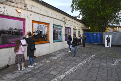 Odwiedzający zgromadzili się na zewnętrznym terenie muzeum kolejnictwa, gdzie na ścianie zawieszono czarno-białe fotografie pociągów i infrastruktury kolejowej. Ludzie rozglądają się i podchodzą do zdjęć, wykazują zainteresowanie eksponatami. Jedna dziewczynka z kilkoma osobami w tle przygląda się fotografii, mając w ręku kartkę i coś pisząc.