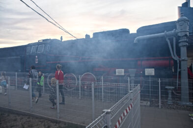 Czarny, parowy lokomotywa emituje gęsty biały dym, podczas gdy liczni odwiedzający przechadzają się wzdłuż ogrodzenia. Niebo ma barwę zmierzchu, a teren wydarzenia jest wyraźnie oznakowany metalowymi barierkami. Widoczne są również torowiska, co wskazuje na kolejowy charakter tego wydarzenia.