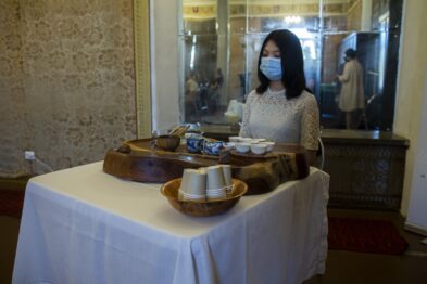 Kobieta stoi przy białym stole, na którym ułożone są eksponaty: ceramiczne naczynia i tradycyjne chińskie instrumenty. Ubrana jest w jasną bluzkę z długimi rękawami i nosi maskę ochronną na twarzy. W tle widać odbicie sali w dużym, ozdobnym lustrze umieszczonym na ścianie.