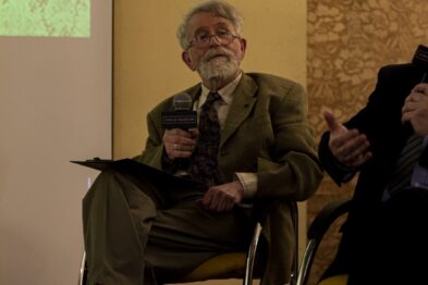 Starszy mężczyzna siedzi na złotej, metalowej krześle ze złożonymi nogami, trzyma mikrofon i mówi, wyrażając emocje gestykulacją rąk. Ubrany jest w beżowy garnitur, białą koszulę i krawat. W tle, nieostrym planie, widać fragment innego uczestnika wydarzenia oraz wyświetlany materiał prezentacyjny.