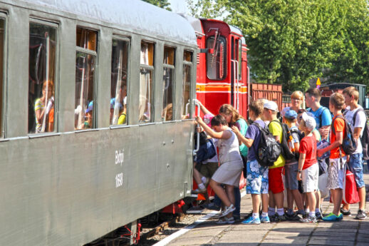 Grupa osób, w tym dzieci i dorośli, stoi na peronie obok starego, zielonego wagonu kolejowego. Czerwony lokomotywa stoi tuż przy nich, a słoneczna pogoda sprawia, że scena jest jasna i barwna. Ludzie są ubrani w letnie stroje, a drzwi wagonu są otwarte, jakby czekano na wejście lub wysiadanie pasażerów.