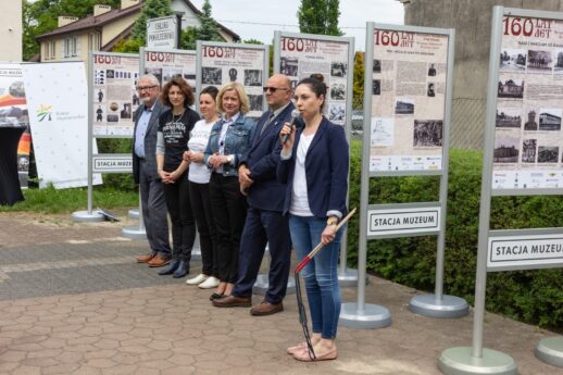 Grupa osób stoi przed stojakami z plakatami, na których wydrukowano informacje i zdjęcia historyczne. W centralnej części widać kobietę trzymającą mikrofon, która wygłasza przemówienie, a obok niej pozostali uczestnicy uroczystości uważnie słuchają. W tle dostrzec można zazieleniony teren i fragmenty budynków.
