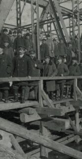 Grupa osób w tradycyjnych ubiorach stoi na drewnianej konstrukcji, będącej zapewne elementem infrastruktury kolejowej. W tle widać wysokie drewniane kratownice, które mogą być częścią mostu lub wiaduktu. Osoby są różnego wieku, a ich postawy sugerują oficjalne pozowanie do zdjęcia.