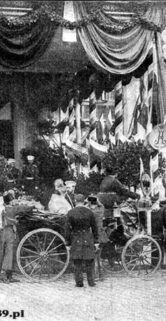 Grupa osób stoi przed ozdobioną fasadą budynku, udekorowanym flagami i tkaninami. Kilka koni przy wozach czeka, a mężczyźni ubrani w ciemne garnitury zgromadzili się wokół. Centralnie w tłumie widoczna jest postać siedząca na wózku inwalidzkim.