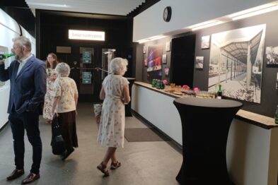 W przestronnej galerii grupa osób ogląda obrazy związane z kolejnictwem eksponowane na ścianach; oświetlenie podkreśla kolory i szczegóły dzieł. Odwiedzający, ubrani w eleganckie stroje, w skupieniu podchodzą do dzieł, niektórzy dyskutują lub przypatrują się z bliska detalom malowideł. W głębi pomieszczenia widać stolik z przekąskami i napojami przygotowanymi dla gości wernisażu.