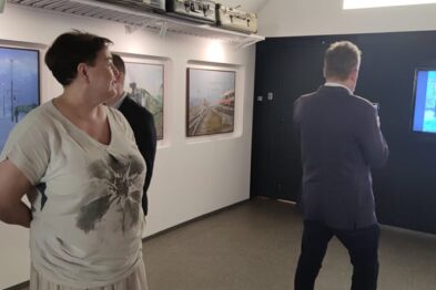 Dwie osoby stoją w pomieszczeniu, gdzie na ścianach zawieszone są obrazy o tematyce kolejowej; jedna z osób ogląda duże płótno z malowidłem pociągu. Na przeciwległej ścianie widać projekcję kolejowego malowidła. W tle oprócz dzieł sztuki widać także elementy wystroju wnętrza, wключая oświetlenie i białe ściany galerii.
