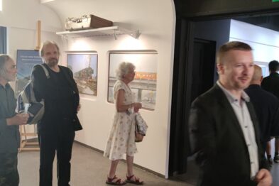 Osoby są ubrane w okolicznościowe stroje i rozmawiają ze sobą, trzymając w rękach kieliszki, co wskazuje na celebrę wydarzenia. Na ścianach widoczne są zawieszone obrazy, a goście przemieszczają się między nimi, przyglądając się dziełom sztuki. Przestrzeń wernisażu wydaje się być jasna i przestronna, oferując komfortowy ogląd ekspozycji.