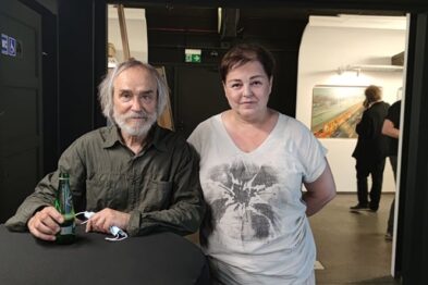 Mężczyzna o siwych włosach i brodzie, ubrany w zieloną koszulę, siedzi przy okrągłym stole trzymając zieloną butelkę i łącząc ręce, na których ma białą maskę. Obok niego stoi kobieta w białej koszulce z nadrukiem, z krótkimi ciemnymi włosami. W tle widać białe ściany galerii oraz kolejne osoby contemplujące obrazy.