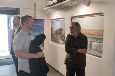 Dwoje osób stoi i rozmawia w galerii, gdzie na ścianach zawieszone są obrazy o tematyce kolejowej. Osoba po prawej stronie ma długie włosy i nosi ciemną kurtkę, trzyma rękę w kieszeni spodni. Na przeciwległej ścianie widać kolejne dzieła sztuki prezentujące pociągi i tory kolejowe.