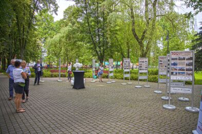 W parku ustawione są stojaki z informacyjnymi banerami, przedstawiającymi historię kolei. Osoby odwiedzające wystawę przeglądają eksponaty oraz czytają zamieszczone treści. Zieleniejące drzewa tworzą przyjemne tło dla wydarzenia kulturalnego na świeżym powietrzu.