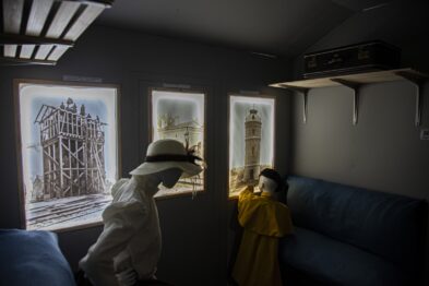 Dwie osoby stoją przed oknem w pomieszczeniu z fotogramami na ścianie, które prezentują konstrukcje kolejowe. Osoba w białym kapeluszu wskazuje palcem na jeden z obrazów, zdaje się opisywać jego zawartość drugiej, młodszej osobie w żółtym płaszczyku. Wnętrze jest oświetlone naturalnym światłem wpadającym przez okno i sztucznym światłem z góry.