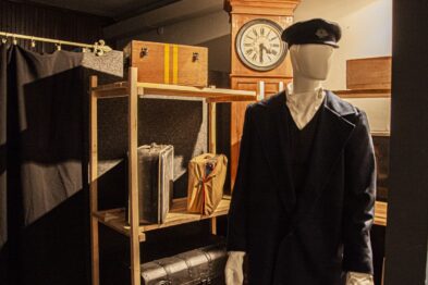 Widoczna jest część ekspozycji muzealnej składająca się z manekina ubranego w ciemny mundur z daszkiem. W tle znajduje się stara walizka oraz drewniana półka z różnymi tekstylnymi eksponatami i zegar ścienny. Całość prezentuje się w tonacji ciemnych barw i jest odpowiednio oświetlona do prezentacji.