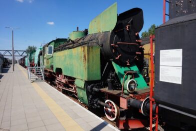 Stary parowóz o zielono-czerwonym malowaniu stoi na torach kolejowych. Obok lokomotywy umieszczona jest tablica informacyjna, a w tle widać kładkę pieszą. Lokomotywa wyposażona jest w tradycyjne elementy takie jak duże koła napędowe, komora kabinowa i dymnica.