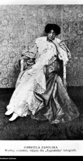 Kobieta w długiej sukni i z dużym kapeluszem przyjmuje elegancką, wyprostowaną pozę. Jest ubrana w sposób, który może sugerować modę z początku XX wieku, ze szczególną uwagą na detale i dodatki takie jak koronki i frędzle. Twarz postaci jest wyraźna i zwrócona w stronę kamery, a tło za nią jest jednolite i dekoracyjne, co dodaje portretowi charakter studyjnej fotografii.