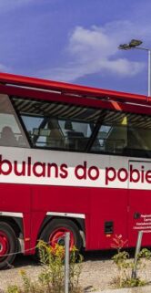 Czerwony ambulans do pobierania krwi stoi zaparkowany na asfaltowym parkingu. Pojazd ma na boku napis "Ambulans do pobierania krwi" i jest wyposażony w sprzęt potrzebny do przeprowadzenia zabiegu. W tle widoczne są drzewa i niebieskie, zachmurzone niebo.