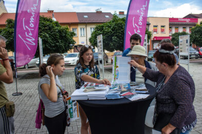 Grupa osób stoi wokół stołu z materiałami promocyjnymi na zewnątrz przy dobrym oświetleniu. Na stole leżą różne broszury i ulotki, a nad stołem znajdują się flagi z napisem 