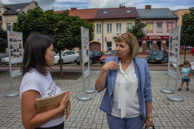 Dwie kobiety rozmawiają na tle plakatów związanych z kolejnictwem. Osoba po prawej gestykuluje podczas rozmowy, ma na sobie niebieską marynarkę i białą bluzkę. W tle widać fragmenty zabudowań miejskich i chodnik.