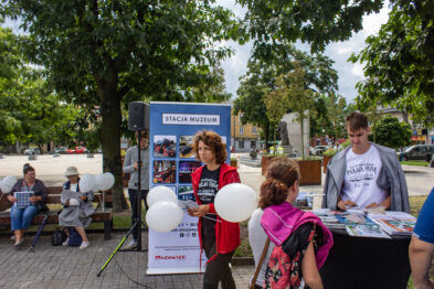 Trwa wydarzenie na świeżym powietrzu z osobami stojącymi przy stolikach z rozdawanymi materiałami. Kobieta w średnim wieku rozmawia z inną osobą; wokół nich balony i plakat z tekstem związanym z historią kolei. Na tle zielonych drzew i nieba z chmurami ludzie siadają na ławkach i spacerują.