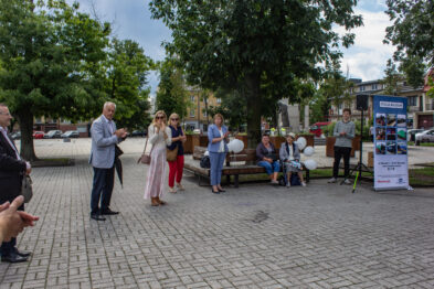 Grupa osób stoi na zewnątrz wokół mówcy z mikrofonem, który wygłasza przemówienie. Widoczne są ekspozycje z informacjami historycznymi o kolei oraz flagi. W środku zgromadzenia jest kobieta w jasnym ubraniu, słuchająca wystąpienia.