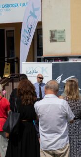 Grupa ludzi stoi na placu przed budynkiem Miejskiego Ośrodka Kultury, gdzie obchodzone jest otwarcie wystawy poświęconej historii kolei. Osoby w różnorodnym ubraniu słuchają mówcy, który stoi obok banera informacyjnego o wydarzeniu. W tle widoczne są plakaty związane z tematyką kolejnictwa oraz flagi.