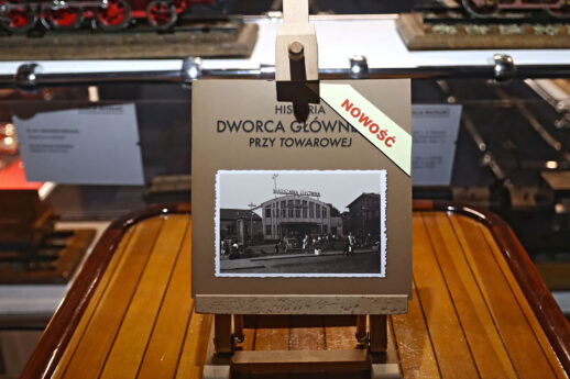 Na drewnianej podstawie stoi album z czarno-białą fotograficzną ilustracją przedstawiającą budynek dworca. Obok albumu widoczne jest oznaczenie 