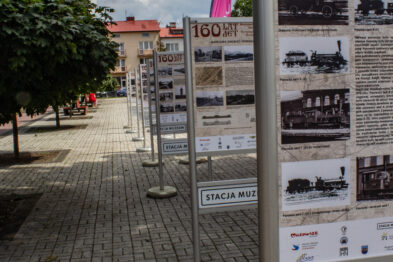 Rząd stojących na bruku informacyjnych banerów przedstawia historię kolei, z jednym wyraźnie zatytułowanym „160 lat Petersbursko – Warszawskiej Drogi Żelaznej”. Fotografia oddaje perspektywę alejki z banerami, które zapraszają do zapoznania się z wystawą. W tle słupki ograniczające przestrzeń i nieliczni przechodnie.