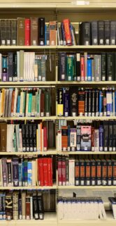 Regały biblioteczne wypełnione książkami o różnej grubości i wysokości tworzą kolorowy mozaik okładek. Kilka książek leży poziomo na półkach. W dolnej części zdjęcia widoczne są etykiety z oznaczeniami systemu klasyfikacyjnego.