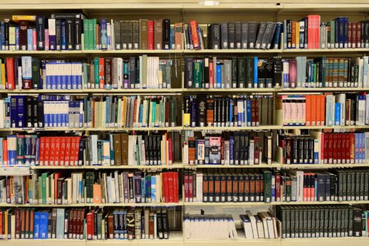 Regały biblioteczne wypełnione książkami o różnej grubości i wysokości tworzą kolorowy mozaik okładek. Kilka książek leży poziomo na półkach. W dolnej części zdjęcia widoczne są etykiety z oznaczeniami systemu klasyfikacyjnego.