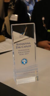 Statuetka o przezroczystej strukturze stoi na stole przed nieostrym tłem, na której wygrawerowane jest imię i nazwisko osoby nagrodzonej oraz tytuł "Mazowiecki Bibliotekarz Roku 2019". W tle widoczne są sylwetki ludzi i fragmenty mebli, co sugeruje miejsce spotkań lub konferencji. Statuetka ma formę prostopadłościanu z wycięciem na górze przypominającym płomień lub kroplę.