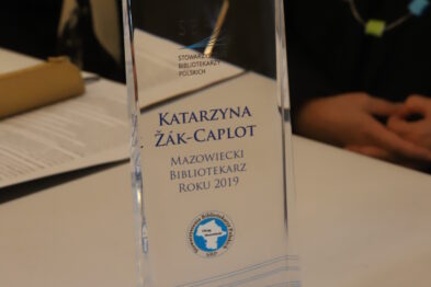 Statuetka o przezroczystej strukturze stoi na stole przed nieostrym tłem, na której wygrawerowane jest imię i nazwisko osoby nagrodzonej oraz tytuł 