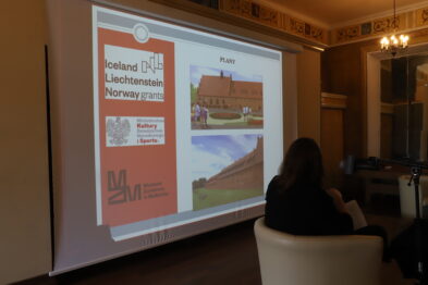 W pomieszczeniu o klasycznym wystroju, z lekkościem nasuwa się atmosfera naukowej wymiany myśli. Widoczny ekran projekcyjny ukazuje prezentację o tematyce bibliotecznej, zaznaczając kontekst kulturowy i historyczny. Osoba siedząca odwrócona tyłem do kamery, z uwagą śledzi wyświetlane treści, podkreślając skupienie i powagę momentu.