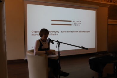 Osoba siedzi na jasnym fotelu i czyta z kartki przed mikrofonem; nad nią wisi projektor z napisem „Organizatorzy: - patronat honorowy Muzeum Kolejnictwa na Śląsku”. W tle widoczne są elementy wnętrza sali z ciemnymi krzesłami oraz ekrany projekcyjne.