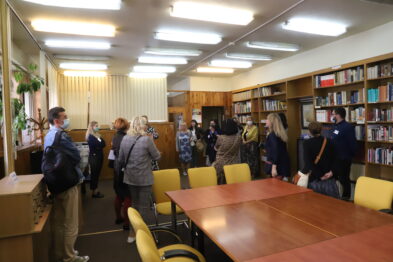 Grupa ludzi stoi w dobrze oświetlonym pomieszczeniu wyposażonym w półki z książkami i stoły z krzesłami. Większość osób skupia się wokół osoby prezentującej coś u góry po lewej stronie. Osoby na zdjęciu noszą maseczki ochronne na twarzach i wydają się uważnie słuchać prelegenta.