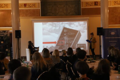 Grupa ludzi siedzi na krzesłach w sali konferencyjnej i ogląda prezentację. Prezenter wskazuje na slajd wyświetlany przez projektor, na którym jest widoczne zdjęcie budynku. Sala jest przestronna z dekoracyjnym sufitem i oknami.