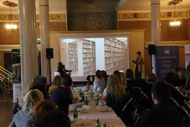 Grupa osób siedzi przy stołach ustawionych w rzędach w jasnym pomieszczeniu; niektórzy uczestnicy konferencji zwracają uwagę na ekran, na którym wyświetlany jest obraz półek z książkami. Prelegentka w środku sali pokazuje na ekran podczas swojej prezentacji, a słuchacze mają przed sobą notatniki i butelki z wodą. Sala jest przestronna, z dekoracyjnym sufitem i dużymi oknami, przez które wpada naturalne światło.