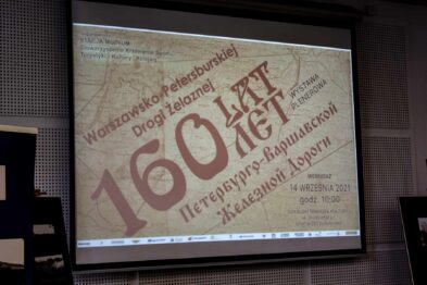 Widoczny jest ekran projekcyjny z wyświetlanym tekstem i grafiką celebrującą 160-lecie istnienia Petersbursko-Warszawskiej Drogi Żelaznej. Na ekranie znajduje się mapa oraz duże, wyraźne napisy, w tym liczba 160, świadczące o rocznicy. Całość jest prezentowana w pomieszczeniu z ciemnym tłem, a ekran stanowi centrum uwagi.
