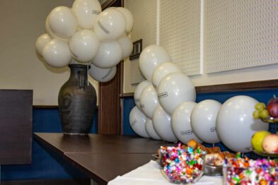 Kompozycja balonów w odcieniach bieli i szarości jest umieszczona w dużym ciemnym wazonie na stole, obok którego znajdują się miski z kolorowymi cukierkami i owocami. Na stole rozłożony jest biały obrus, a w tle widoczna jest jasna ściana z roletą okienną. Całość tworzy dekorację celebrującą wydarzenie.