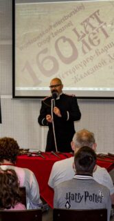 W pomieszczeniu z zebranymi ludźmi mężczyzna stoi na przodzie i przemawia do publiczności; w tle widoczne są plakaty związane z kolejnictwem. Uczestnicy siedzą na krzesłach i słuchają prelegenta; niektórzy z nich zwracają uwagę na fotografa. Pomieszczenie ma duże okna i eleganckie, jasne ściany, a wystawa zdaje się mieć charakter edukacyjny.