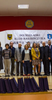 Grupa osób stoi na scenie przed szerokim banerem z logo Dolnośląskiego Klubu Harbińczyka i herbem miasta. Ludzie pozują do zdjęcia, są uśmiechnięci, ubrani w eleganckie stroje. W tle widać dekoracje kwiatowe oraz sprzęt nagłaśniający.