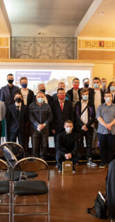 Grupa osób stoi do zdjęcia w przestronnej sali, w której widać ustawione rzędy krzeseł i dekoracje na ścianach. Część osób ma na twarzach maski ochronne, a ich stroje wskazują na formalne lub półformalne wydarzenie. W tle znajduje się ekran z projekcją oraz liczne materiały edukacyjne związane z tematyką kolejnictwa.