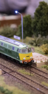Na pierwszym planie znajduje się zielony model pociągu poruszający się po miniaturowych torach kolejowych; otacza go realistycznie odwzorowana roślinność i infrastruktura kolejowa. Model ten jest częścią precyzyjnie wykonanej makiety kolei, która symuluje rzeczywiste otoczenie torowisk z uwzględnieniem szczegółów takich jak podkłady i żwir. W tle widoczne są kolejne elementy ekspozycji, które dopełniają całości wyglądu miniaturow