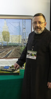 Mężczyzna w ciemnym ubiorze prezentuje modele kolejowe wystawione na zielonym stole. Za nim dwie duże plakietki z widokami krajobrazów kolejowych w tle. Na stoisku znajdują się również ulotki i inne materiały promocyjne.