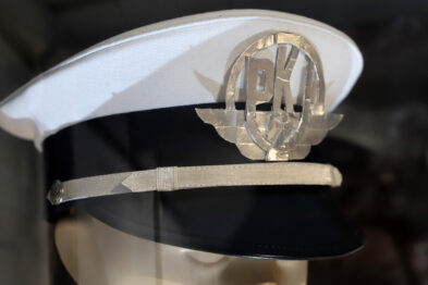 Biało-czarna czapka kolejarska z czarnym daszkiem i ozdobnym emblematem umieszczonym na przodzie. Emblemat przedstawia skrzyżowane młotki z zaokrąglonymi kształtami w tle, które mogą symbolizować koła lub elementy techniczne. Czapka ma lśniącą czarną opaskę oraz srebrną lamówkę przy daszku.