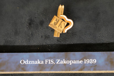 Złota odznaka z napisem „FIS” jest wyeksponowana na ciemnym tle, spoczywa na czarnej powierzchni z opisem w jasnej czcionce. Odznaka ma formę skrzyżowanych nart z pięcioma pierścieniami olimpijskimi i inicjałami „FIS”. Pod spodem na etykiecie napisano „Odznaka FIS, Zakopane 1939”.