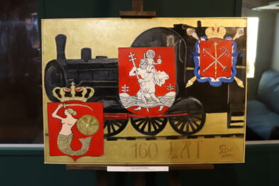 Na obrazie widoczna jest lokomotywa parowa umiejscowiona pośrodku, ozdobiona herbami po bokach. Po lewej stronie lokomotywy umieszczono czerwony herb z białym orłem, a po prawej - herb z czerwonym rycerzem na białym tle. Dół obrazu zdobi złoty napis z datą 